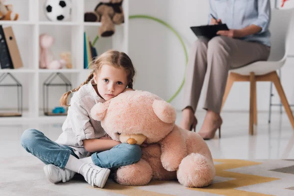 Сумна маленька дитина з плюшевим ведмедем сидить на підлозі з психологом, сидячи на фоні — стокове фото