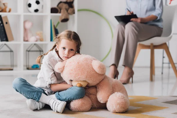 Triste niño pequeño con osito de peluche sentado en el suelo con psicólogo sentado en el fondo - foto de stock