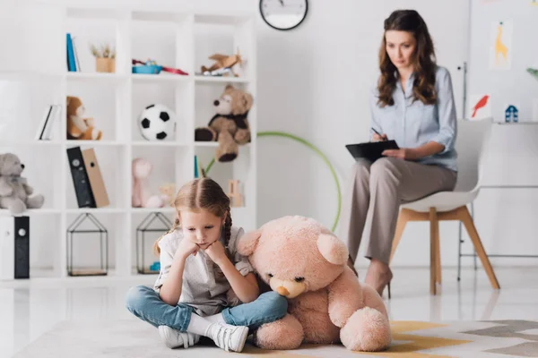 Niedergeschlagenes kleines Kind mit Teddybär auf dem Boden sitzend mit verschwommenem Psychologen im Hintergrund — Stockfoto