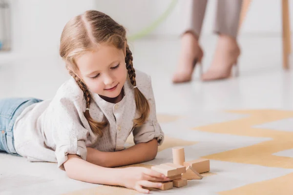 Маленька дитина грає з дерев'яними блоками на підлозі з розмитим психологом, сидячи на фоні — стокове фото