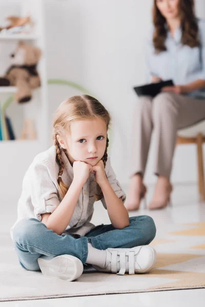 Подавленный маленький ребенок смотрит в камеру, сидя на полу с размытым психологом на заднем плане — стоковое фото