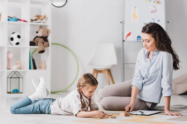 Psychologue souriant assis près de l'enfant alors qu'elle est couchée sur le sol et jouer avec des blocs de bois — Photo de stock