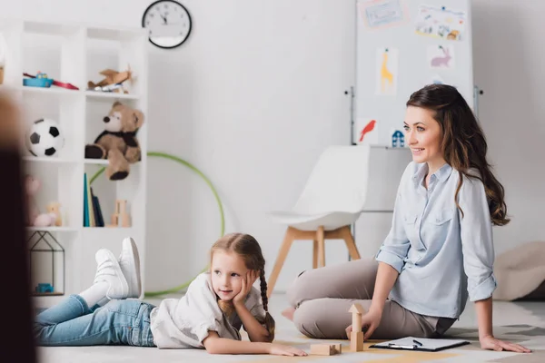 Sorridente psicologa seduta vicino al bambino mentre si sdraia sul pavimento e distoglie lo sguardo — Foto stock