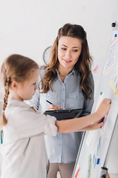 Psicologo sorridente con appunti e bambino vicino alla lavagna bianca con vari disegni — Foto stock