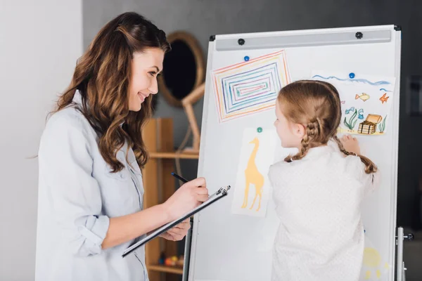 Happy psychologist with clipboard talking to child near whiteboard with various drawings — Stock Photo