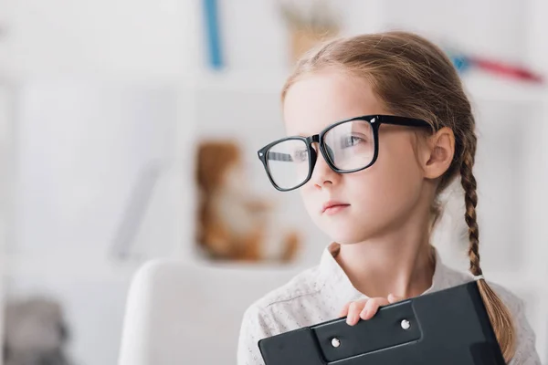 Nahaufnahme Porträt eines kleinen Kindes mit Brille, das Klemmbrett hält und wegschaut — Stockfoto