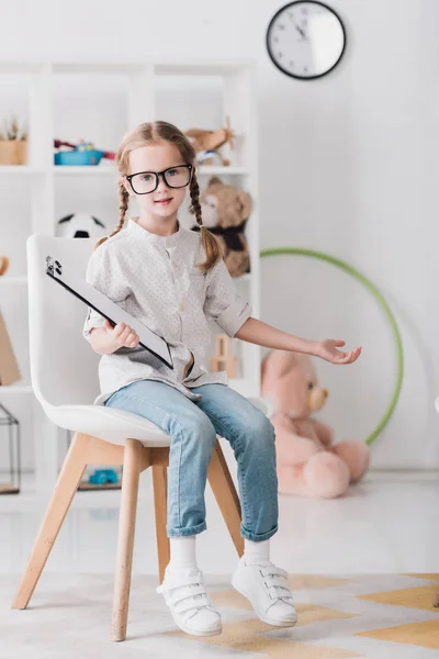 Маленький ребенок в очках с планшетом сидит на стуле и смотрит в камеру — стоковое фото