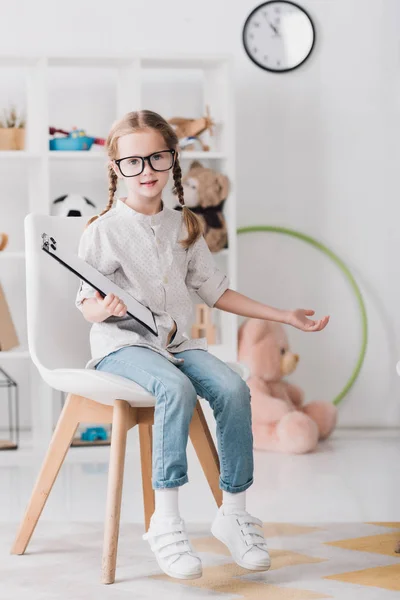 Niño pequeño en gafas con portapapeles sentado en la silla y mirando a la cámara - foto de stock