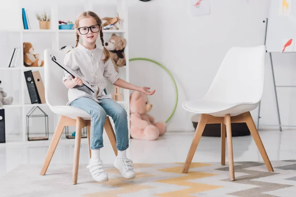 Adorable little child in eyeglasses with clipboard sitting on chair and looking at camera — Stock Photo