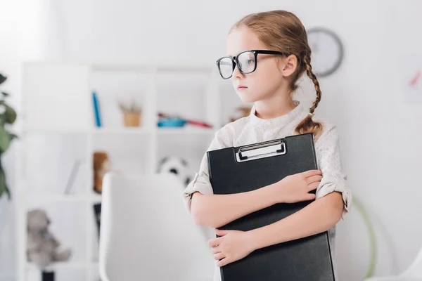 Sérieux petit enfant dans les lunettes tenant presse-papiers et détournant les yeux — Photo de stock