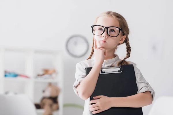 Petit enfant réfléchi dans des lunettes avec presse-papiers regardant vers le haut — Photo de stock