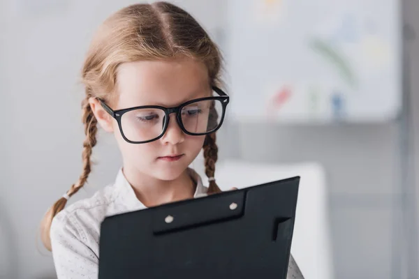 Nahaufnahme Porträt eines ernsthaften kleinen Kindes mit Brille, das im Klemmbrett schreibt — Stockfoto