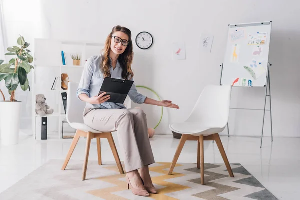 Psicólogo infantil feliz con portapapeles apuntando al asiento y mirando a la cámara — Stock Photo