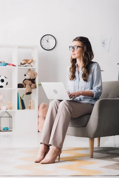 Child psychologist sitting in armchair with laptop and looking away — Stock Photo