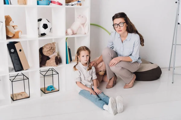 Psicóloga mujer apoyando a un niño deprimido mientras se sienta en el suelo y mira a la cámara - foto de stock