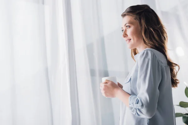 Happy adult woman holding cup of warming beverage and looking away — Stock Photo