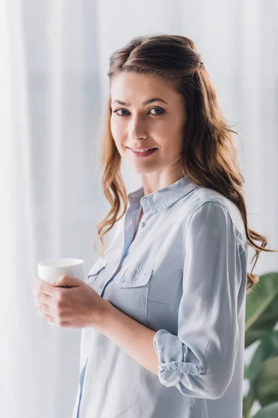 Счастливая взрослая женщина держит чашку согревающего напитка и смотрит в камеру — стоковое фото