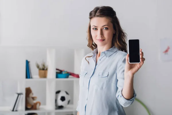 Femme adulte souriante en chemise montrant smartphone avec écran blanc à la caméra — Photo de stock