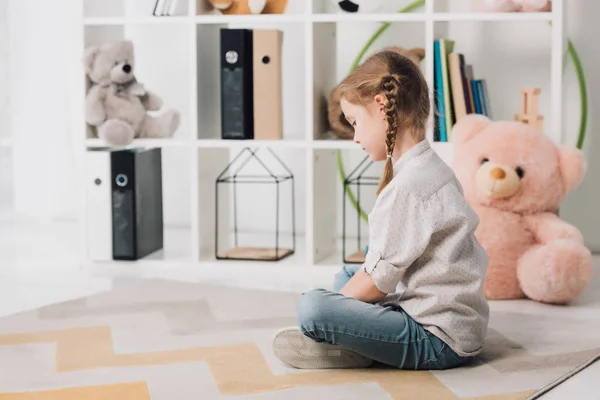 Lonely little child sitting on floor in front of shelves with toys — Stock Photo