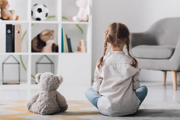 Rear view of little child in white shirt sitting on floor with teddy bear — Stock Photo