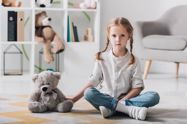 Petit enfant solitaire assis sur le sol avec un ours en peluche et regardant la caméra — Photo de stock