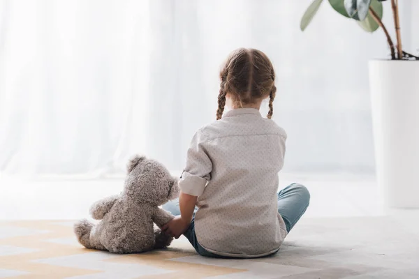 Rear view of little child sitting on floor with her teddy bear toy — Stock Photo