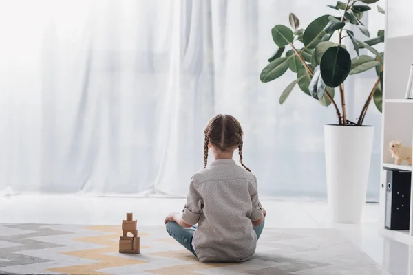 Vista trasera del niño pequeño sentado en el suelo con bloques de madera y mirando hacia otro lado - foto de stock