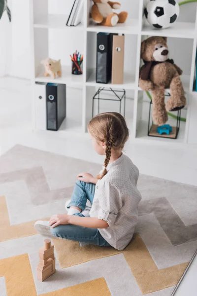 Vista de ángulo alto del niño pequeño deprimido sentado en el suelo con bloques de madera y mirando hacia otro lado - foto de stock