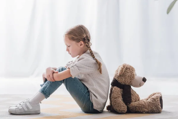 Vue latérale du triste petit enfant assis sur le sol dos à dos avec ours en peluche — Photo de stock