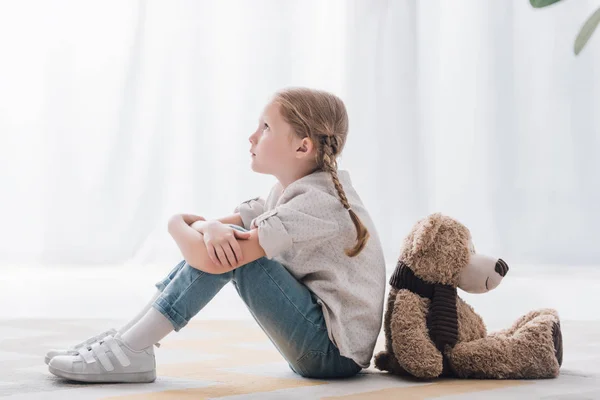 Vue latérale du petit enfant réfléchi assis sur le sol dos à dos avec ours en peluche — Photo de stock