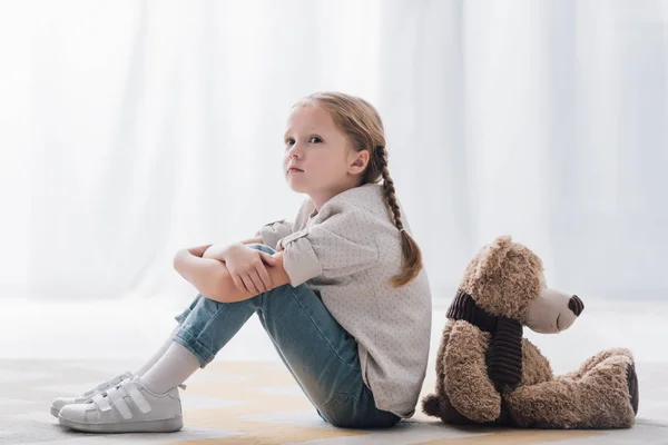 Side view of depressed little child sitting on floor back to back with teddy bear — Stock Photo