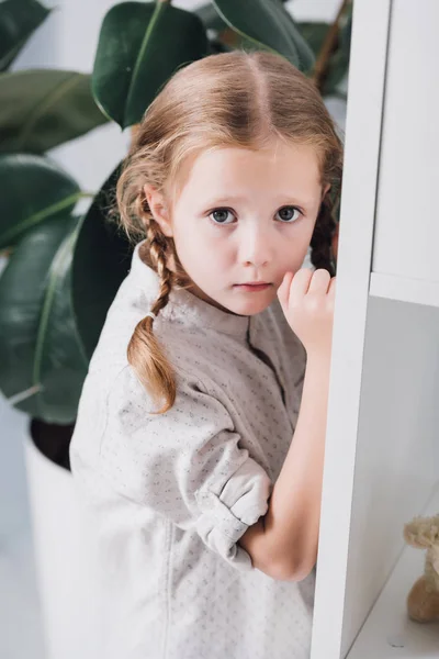 Scared little child standing behind bookshelves and looking at camera — Stock Photo