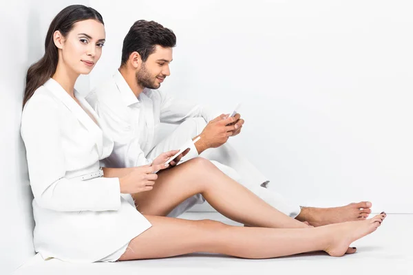 Belle femme en blanc total en utilisant une tablette numérique tout en tapant l'homme sur smartphone — Photo de stock