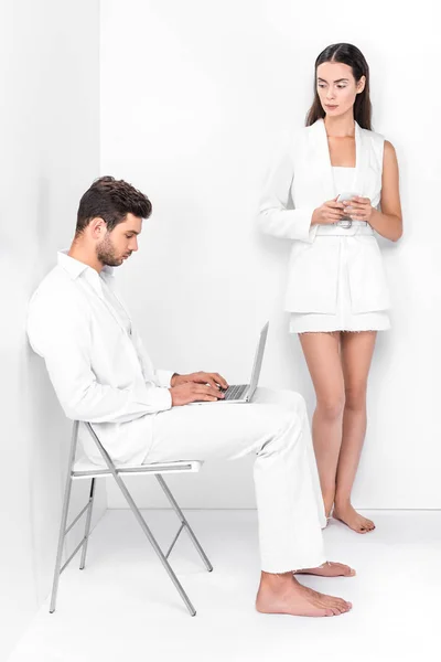 Hombre adulto en blanco total usando el ordenador portátil, mientras que la mujer elegante escribiendo en el teléfono inteligente - foto de stock