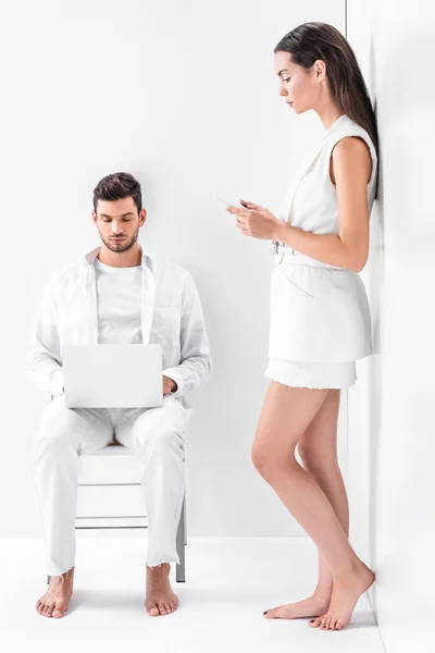 Hombre adulto en blanco total sentado en la silla y escribir en el ordenador portátil, mientras que la mujer atractiva utilizando el teléfono inteligente - foto de stock