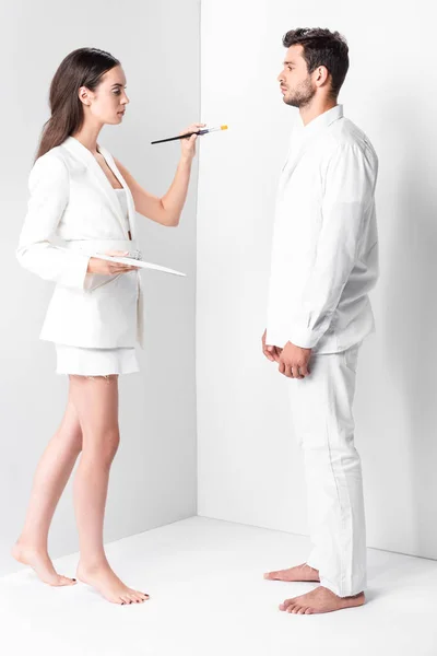Erwachsene Künstlerin in ganz weißer Haltung mit Palette und Pinsel vor dem Mann — Stockfoto