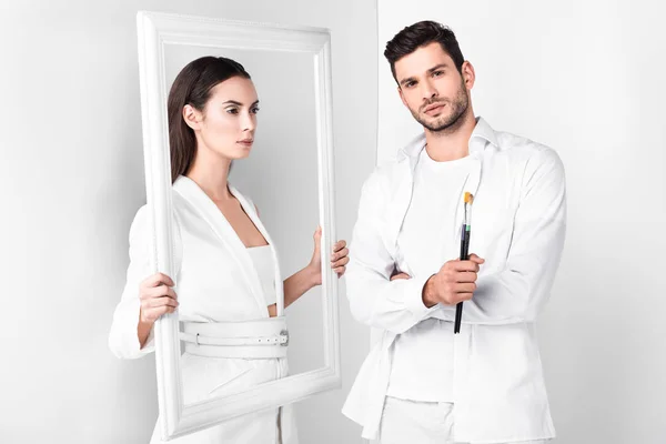 Attractive adult woman holding frame near man with paintbrush on white background — Stock Photo