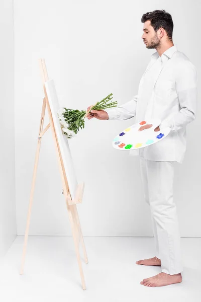 Barefoot male artist in total white drawing on easel with bouquet of flowers — Stock Photo