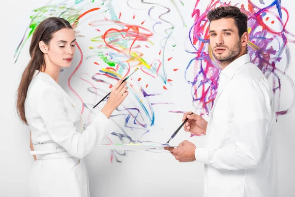 Close up of adult couple in total white drawing with paintbrushes together on wall — Stock Photo