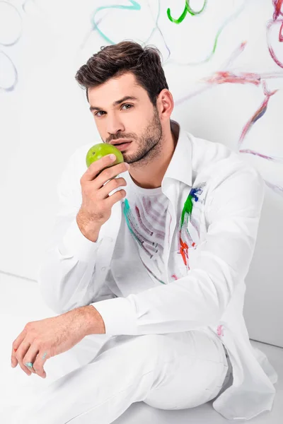 Close up of adult man in total white with ripe apple sitting near painted wall — Stock Photo