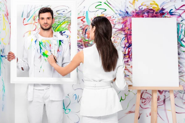 Schöne erwachsene Frau zeichnet auf Kleidung, während Mann Rahmen in der Nähe bemalter Wand hält — Stockfoto