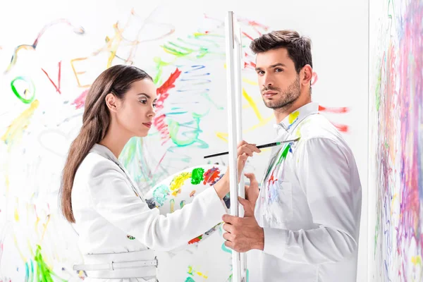 Close up of adult man holding white frame while woman painting clothes — Stock Photo