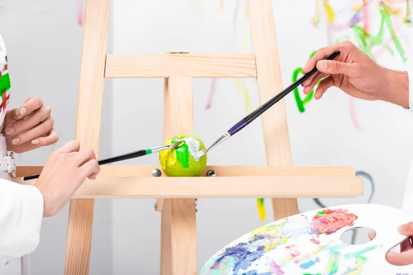 Close up of adult couple painting green apple on easel with paintbrushes — Stock Photo