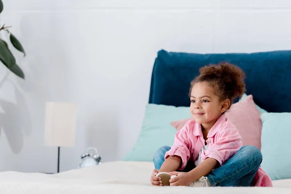 Lächelnd afrikanisches amerikanisches Kind sitzt auf dem Bett und umklammert ihre Beine — Stockfoto