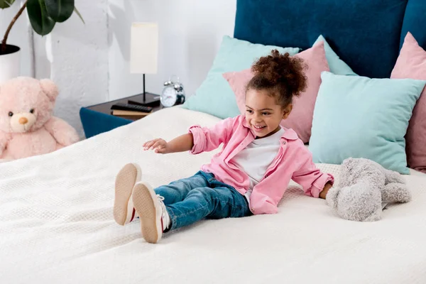 Juguetón afroamericano niño en la cama con osito de peluche - foto de stock