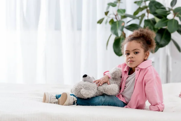 Niña afroamericana trastornada sentada en la cama con oso de peluche de rodillas - foto de stock