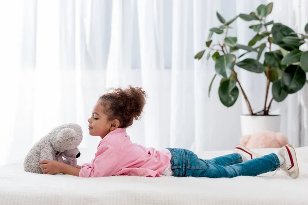 Vista lateral del niño afroamericano acostado en la cama y jugando con su osito de peluche - foto de stock