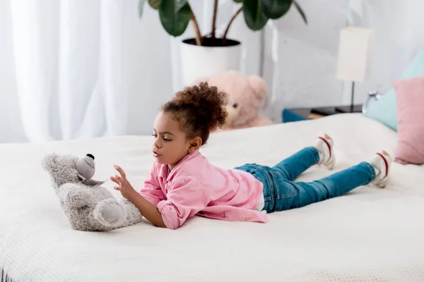 Adorável encaracolado afro-americano criança deitada na cama e brincando com seu urso de brinquedo — Fotografia de Stock