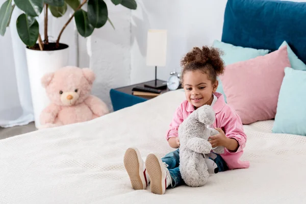 Sonriente niña afroamericana sentada en la cama y jugando con su osito de peluche - foto de stock