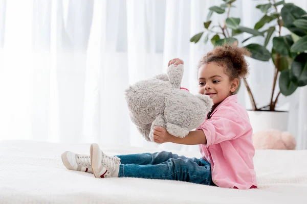 Süße afrikanische amerikanische Kind sitzt auf dem Bett und spielt mit ihrem Teddybär — Stockfoto